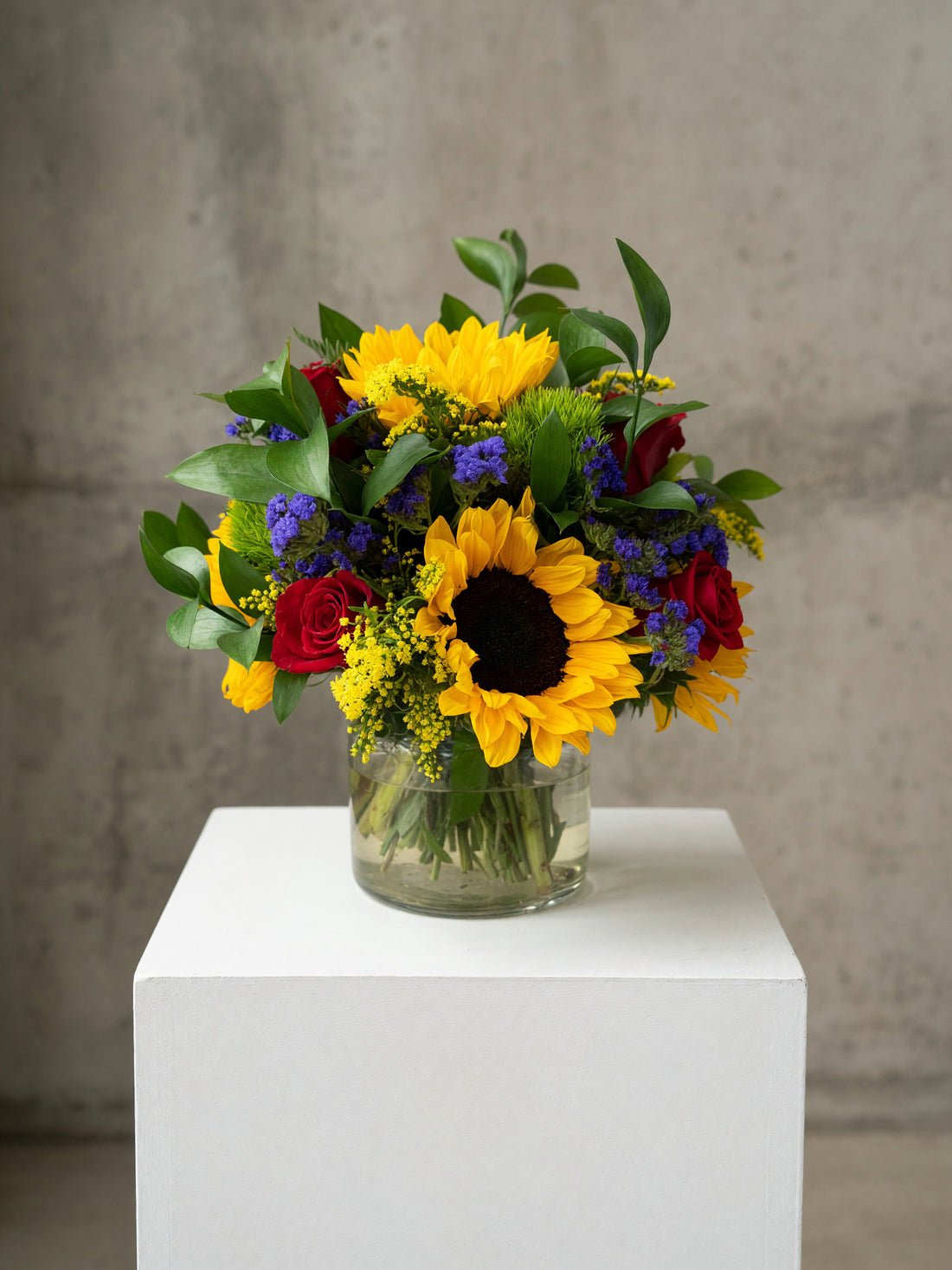 A vibrant mixed flower bouquet featuring a large yellow sunflower, red roses, purple flowers, and green foliage, arranged in a clear glass jar vase and displayed on a white pedestal against a textured gray concrete wall.