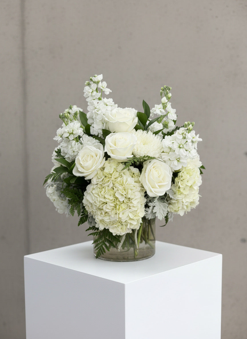 A white floral arrangement featuring roses, hydrangeas, and Stock in a clear glass vase, displayed on a white pedestal against a neutral gray wall.