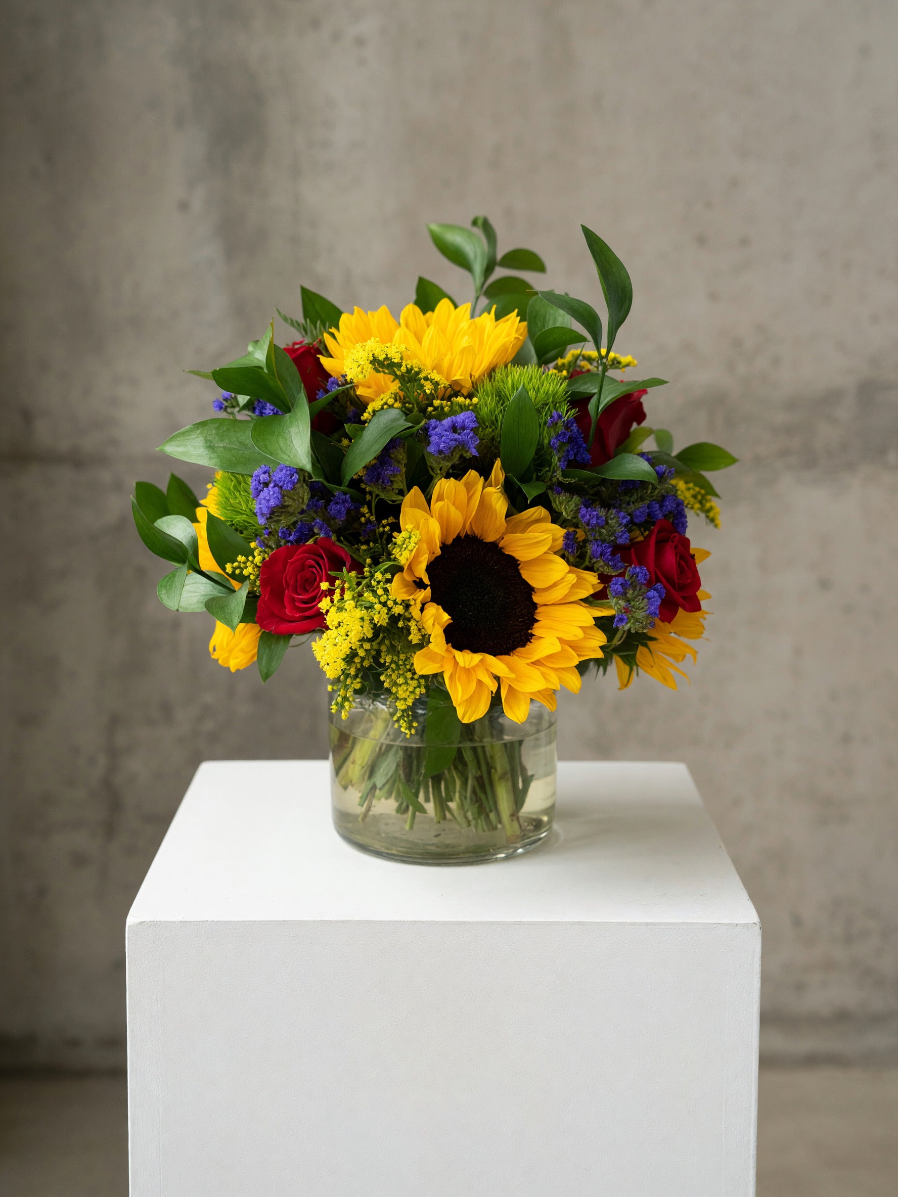 A vibrant mixed flower bouquet featuring a large yellow sunflower, red roses, purple flowers, and green foliage, arranged in a clear glass jar vase and displayed on a white pedestal against a textured gray concrete wall.