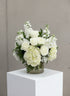 A white floral arrangement featuring roses, hydrangeas, and Stock in a clear glass vase, displayed on a white pedestal against a neutral gray wall.